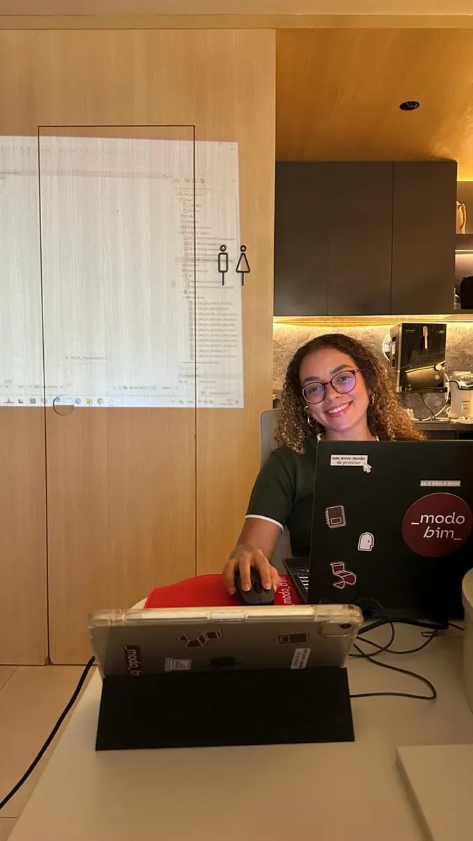 Aluna sorrindo à mesa, com o notebook coberto de adesivos da _modo_bim e uma tela de projeto BIM projetada na parede atrás